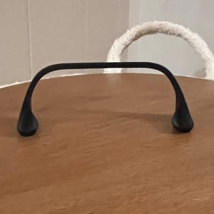 Op de afbeelding: Een zwarte metalen handgreep met druppelvormige uiteinden is gecentreerd op een houten oppervlak. De handgreep buigt omhoog. Op de achtergrond een witte touwhandgreep en een neutraal gekleurde muur.