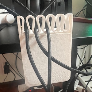 Könnte beinhalten: Ein weißer, 3D-gedruckter Kabelorganisator mit strukturierter Oberfläche und Schlaufenhaltern. Schwarze Kabel sind in den Organizer eingesetzt, im Hintergrund ist ein schwarzer Kopfhörer zu sehen. Der Organizer steht auf einem schwarzen Schreibtisch.