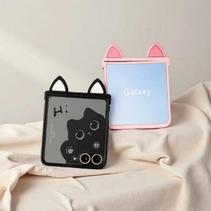 May include: Two phone cases with cat ear designs. One case is black with a cartoon cat graphic and white ears. The other is pink with pink ears and the word "Galaxy" on the screen. Both cases are on a beige fabric.