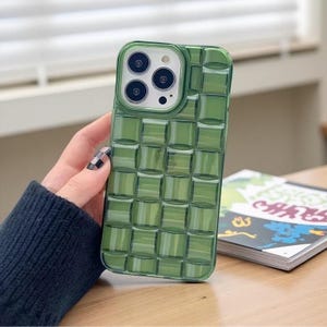May include: A green phone case with a woven pattern. The case has a square grid design and a clear, transparent appearance. The phone case is held by a person with a dark blue sleeve and a black and white checkered nail.