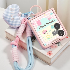 Könnte beinhalten: Eine rosa und blaue Handyhülle mit passendem Lanyard und herzförmigem Anhänger. Die Handyhülle hat ein Cartoon-Design mit dem Text "Pink house". Das Lanyard ist rosa und blau.