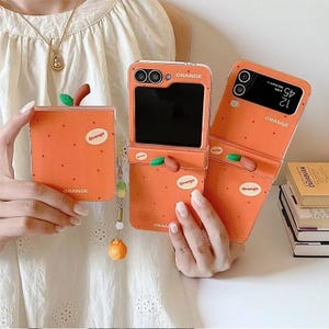 May include: Orange-themed phone cases with a fruit design. The cases are orange with the word "Orange" printed on them. One case is closed, and two are open, displaying the phone screens. Each case has a small, green leaf and brown stem accent.