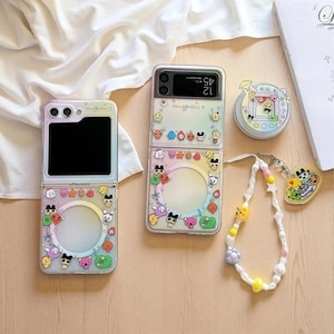May include: Clear phone cases with cartoon character designs. The cases are for a foldable phone, with a circular cutout and a rainbow border. A beaded charm necklace and a round container are also present.