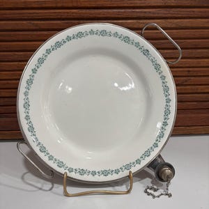 Puede incluir: Plato de cerámica blanca con borde plateado y delicado borde floral verde. El plato se exhibe sobre un soporte metálico con asa y boquilla decorativa con cadena.