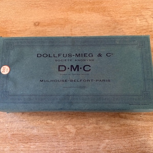 Puede incluir: Una caja rectangular turquesa con el texto "DOLLFUS-MIEG & C", "D.M.C" y "MULHOUSE-BELFORT-PARIS". La caja tiene un borde decorativo y una pequeña pegatina circular. Descansa sobre una superficie de madera.