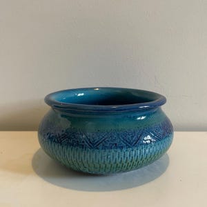 May include: A round ceramic bowl featuring a glossy finish with a gradient of deep blue at the rim transitioning to a lighter teal and green at the base. The exterior surface is decorated with a textured geometric pattern of chevrons and vertical lines.