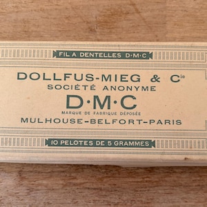 May include: A rectangular box of DMC thread, with text in French. The box is beige with green text and decorative borders. The text includes "FIL A DENTELLES D.M.C" and "10 PELOTES DE 5 GRAMMES". A red circle with "Art. 19" is on the left.