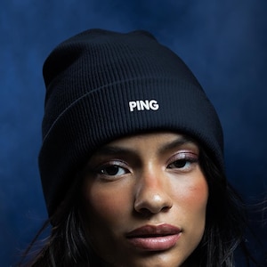 May include: A black ribbed knit beanie with the word PING embroidered in white on the front, worn with a cream-colored fleece zip-up jacket, a black quilted winter coat, and white fingerless knit gloves.