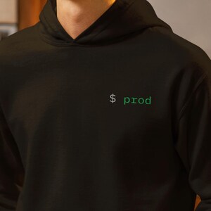 May include: Black hoodie with the text "$ prod" in green. The hoodie has a front pocket and a drawstring hood. The person in the image is holding a gray mug. The background is a kitchen setting.