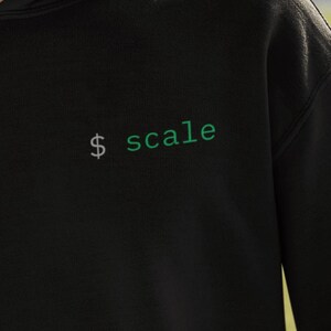 May include: Black hooded sweatshirt with the word "scale" in green and a dollar sign. The sweatshirt is made of a soft material and has a drawstring hood and a front pocket. The person in the image is wearing the sweatshirt.