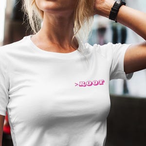 May include: White t-shirt with the word ">ROOT" in pink, small text on the upper chest. The shirt is paired with blue jeans. The model is standing outside.