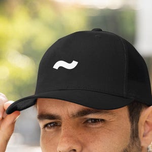 May include: Black baseball cap with a white curved line design. The cap has a curved brim and a mesh back. The person in the image is wearing a navy blue quilted jacket.
