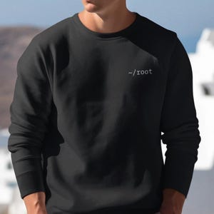 May include: A black long-sleeve crewneck sweatshirt featuring the text -/root printed in white on the upper left chest. The garment has a ribbed neckline, cuffs, and hem, and is paired with tan shorts and round sunglasses.