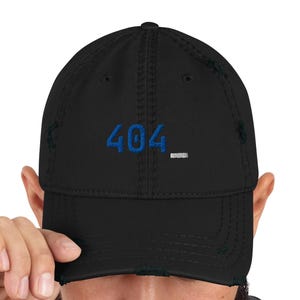 May include: A black distressed baseball cap featuring the embroidered blue text 404 followed by a white underscore symbol. The cap has a structured crown, a curved brim with frayed edges, and matching black stitching throughout the design.