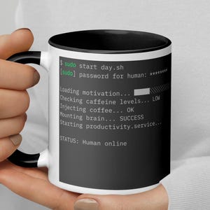May include: A white coffee mug with a black interior and handle, featuring text in green and white. The text simulates a computer terminal's startup sequence, including "Loading motivation..." and "STATUS: Human online."