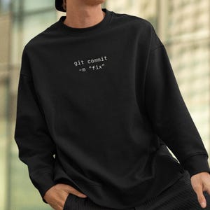 May include: A black long-sleeve sweatshirt featuring the white text "git commit -m "fix"" printed on the chest. The garment has a crew neckline, ribbed cuffs, and a relaxed fit, constructed from a solid black fabric with clean, minimalist lines.