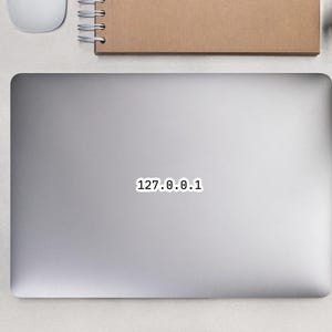 May include: A laptop with a sticker that reads "127.0.0.1" is on a light gray surface. A white computer mouse, a spiral-bound notebook, a pencil, glasses, and small potted plants are also on the surface.