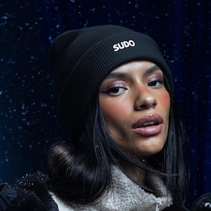 May include: A black ribbed knit beanie featuring the word SUDO embroidered in white lettering on the front cuff. The beanie is worn with a cream-colored fleece zip-up jacket layered under a black quilted winter coat with silver metal snaps.