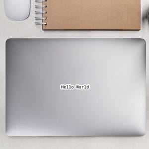 May include: A laptop with the text "Hello World" on the screen, a spiral-bound notebook, a white mouse, a black pencil, a pair of black glasses, and two small potted plants are arranged on a light gray surface.
