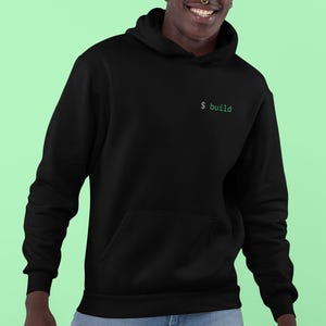 May include: Black hoodie with a front pocket and drawstring hood. The text "$ build" is printed in green on the upper left chest. The hoodie is worn by a person wearing blue jeans.