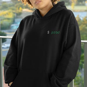 May include: Black hoodie with a front pocket and a hood. The hoodie has the text "$ prod" in green on the upper chest. The person in the image is wearing sunglasses and gold hoop earrings.