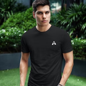 May include: Black crew neck t-shirt with a white "A" logo on the chest. The shirt is paired with blue jeans. The person is wearing a watch and bracelet. The background features greenery and a building.