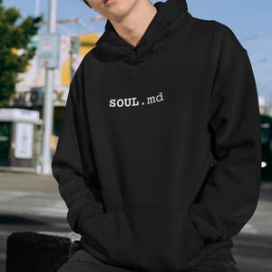 May include: A black hooded sweatshirt featuring the white text SOUL.md printed on the center of the chest. The garment includes a front kangaroo pocket, long sleeves, and a hood with a drawstring. The fabric has a smooth, solid black finish.