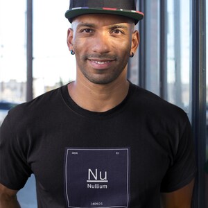 May include: A black t-shirt featuring a white graphic square resembling a periodic table element with the text 404, Er, Nu, Nullium, and 404.0. The shirt is paired with a black baseball cap that has a multicolored patterned brim and blue hoop earrings.