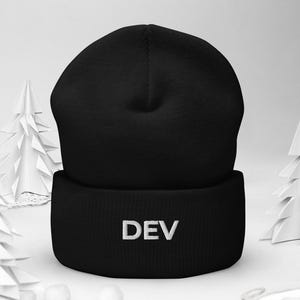 May include: Black beanie hat with the word "DEV" embroidered in white. The hat is on a white surface with paper trees and small white spheres.