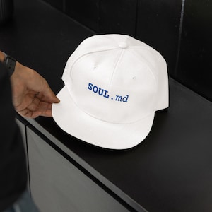 May include: A white baseball cap featuring the text SOUL.md embroidered in blue thread on the front panel. The cap has a structured crown, a flat brim, and a button at the top centre. The design is clean and simple with a bold colour contrast.