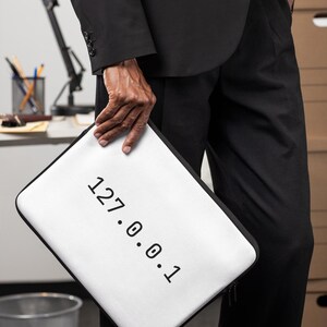 May include: A white rectangular laptop sleeve with black trim and the text 127.0.0.1 printed in black on the front. The sleeve features a zipper closure along the side and is held in a vertical orientation against a dark suit and trousers.