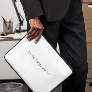 May include: A white rectangular laptop sleeve with black trim and a zip closure. The front of the sleeve features the black printed text $ echo "hello world" in a monospaced font, centred on the white surface.