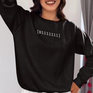 Puede incluir: Una sudadera negra de manga larga con cuello redondo y pu&ntilde;os acanalados. En el centro del pecho aparece el texto blanco [01111111] en una fuente sencilla sin remates. La prenda se combina con una parte inferior de color blanco.