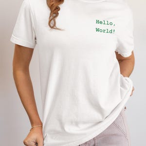 May include: White t-shirt with the text "Hello, World!" in green. The shirt is a classic crew neck style. The model is wearing light pink pants.
