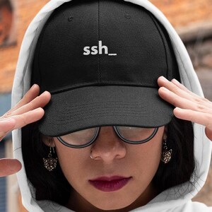 May include: A black baseball cap featuring the white embroidered text "ssh_" on the front panel. The cap has a structured crown and a curved brim, constructed from a solid black fabric material with matching stitching details throughout the design.