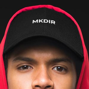 Peut inclure: Un sweat-shirt à capuche rouge vif avec un cordon de serrage et une casquette de baseball noire portant le texte blanc MKDIR brodé sur le panneau avant. Le sweat-shirt a une texture lisse et un motif géométrique subtil sur le côté droit.