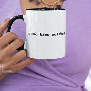 May include: A white ceramic mug with a black handle and interior. The exterior features the black text "sudo brew coffee" printed in a typewriter-style font. The mug is cylindrical with a smooth finish and a standard C-shaped handle.