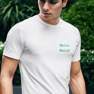 May include: A white short-sleeved t-shirt featuring the text "Hello, World!" printed in green on the left chest area. The shirt has a crew neckline and a standard fit, paired with blue denim jeans that have a slightly distressed texture.