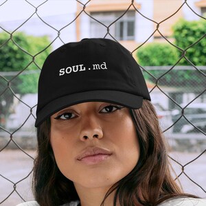 May include: A black baseball cap featuring the white embroidered text SOUL.md on the front panel, paired with a plain white short-sleeved t-shirt. The cap has a curved brim and a structured crown, while the t-shirt has a classic crew neckline.