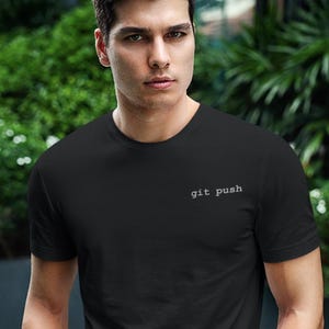 May include: Black crew neck t-shirt with the text "git push" in a small, white font. The shirt is worn with blue jeans. The person is holding sunglasses.