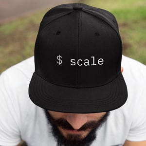 May include: Black baseball cap with the word "scale" in white, featuring a dollar sign. The cap is a flat-brim style, and the text is centered on the front. The cap is worn by a person.