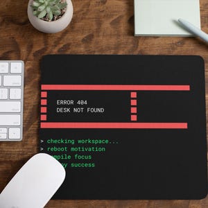 May include: Black mousepad with the text "ERROR 404 DESK NOT FOUND" in white and red. The mousepad also has the text "checking workspace...", "reboot motivation", "compile focus", and "y success" in green. A white keyboard and mouse are also visible.