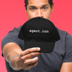 May include: Black baseball cap with the text "agent.run" in white. The cap is held up in front of a person against a red background. The cap is a classic six-panel design.