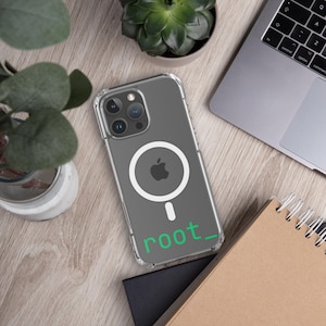 May include: Clear iPhone case with a white MagSafe circle and Apple logo. The case has the word "root_" printed in green. The phone case is on a wooden surface with a laptop, notebook, and plants.