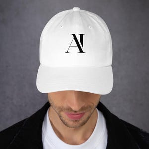 May include: A white baseball cap with a curved brim. The cap features a black stylized "A" and "N" design on the front. The hat is made of a soft material and is suitable for everyday wear.