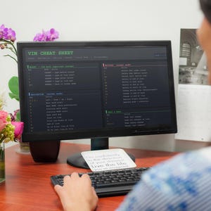 Pu&ograve; includere: Un monitor di computer che mostra una "VIM CHEAT SHEET" con frammenti di codice. Una tastiera e un post-it con testo sono di fronte al monitor. Un vaso di fiori &egrave; a sinistra. La scena &egrave; ambientata su una scrivania di legno.