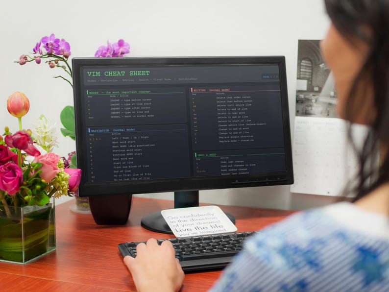 Pu&ograve; includere: Un monitor di computer che mostra una "VIM CHEAT SHEET" con frammenti di codice. Una tastiera e un post-it con testo sono di fronte al monitor. Un vaso di fiori &egrave; a sinistra. La scena &egrave; ambientata su una scrivania di legno.