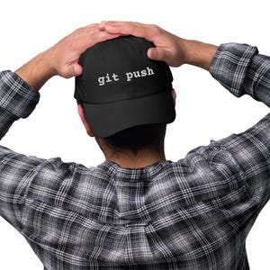 May include: Black baseball cap with the text "git push" in white. The cap is worn by a person wearing a black and white plaid shirt.