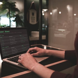 Pu&ograve; includere: Un laptop mostra una "Cheat Sheet" sullo schermo. Il testo verde risalta su uno sfondo scuro. Il laptop &egrave; appoggiato su una superficie di legno scuro. Le mani della persona sono sulla tastiera.