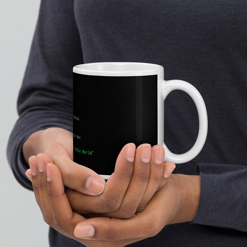 May include: A white ceramic mug featuring a black background with green computer terminal text. The visible text includes status, dev, and the phrase "Hello World". The mug has a standard handle and a smooth, cylindrical body design.